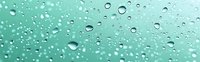 Water drops on turquoise glass surface create natural pattern of scattered transparent spheres with light reflections and bokeh effect on mint background.