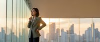 Professional Asian businesswoman talking on phone in modern office with city skyline view during golden hour sunset for corporate communication concepts.
