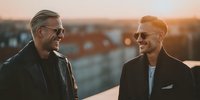 Two professional businessmen in suits having friendly conversation on urban rooftop during golden hour sunset, representing partnership and success.