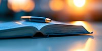 Open book with pen lying across pages against blurred warm lights background, creating atmospheric study and writing composition in blue tones.