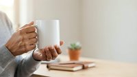 Unrecognizable person with medium skin tone holding blank white mug at wooden desk with books and potted plant, suitable for branding and marketing mockups.