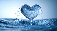 Heart shaped water splash emerging from blue rippling surface with droplets frozen in motion, creating romantic aqua sculpture against soft blue background.