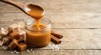 Golden caramel sauce dripping from wooden spoon into glass jar with toffee candies and cinnamon sticks on rustic wood background for dessert recipes.
