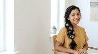 Young South Asian woman with long black hair adorned with white flowers, wearing mustard yellow top, smiling while seated indoors with bright natural lighting.