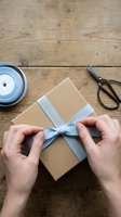 Hands tying light blue ribbon bow on kraft paper gift box on wooden surface with scissors and ribbon spool nearby, overhead view for wrapping and packaging projects.
