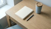 Blank notebook with pen and coffee cup on wooden desk near window, minimalist workspace setup for creative work and planning.