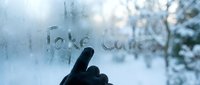 Gloved hand writing Take Care on a foggy winter window with snowy trees in the background, evoking warmth and care.