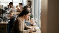 Young Asian woman in brown sweater sitting alone at cafe table by window, contemplating while holding coffee cup in cozy coffee shop atmosphere.