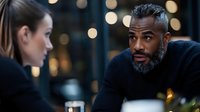 Mature African American businessman with gray beard in discussion with female colleague at evening meeting, bokeh lights background creates professional atmosphere.