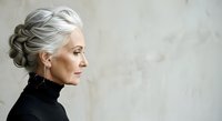 Mature woman with silver gray hair styled in elegant updo, wearing black turtleneck, profile view against neutral textured background. Suitable for beauty, aging gracefully, and lifestyle content.