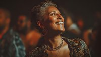 Mature woman with silver hair laughing joyfully at cultural event, wearing ornate patterned outfit and gold jewelry against dark background.