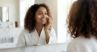 Young African American woman applying skincare cream in bathroom mirror, morning beauty routine, natural curly hair, white bathrobe, self-care wellness lifestyle.