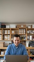 Smiling middle-aged Caucasian businessman working on laptop in organized home office with wooden shelves and storage boxes for small business operations.