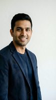 Confident South Asian businessman in navy blazer smiling at camera against neutral background for corporate headshot and professional marketing materials.