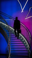 Silhouetted male professional ascending modern staircase with briefcase against vibrant neon blue and purple gradient background with geometric lighting elements.