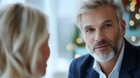 Mature businessman with gray beard and stylish haircut in navy suit smiling confidently during meeting with female colleague, blurred bokeh background.