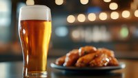 Cold glass of draft beer with foam head next to plate of buffalo chicken wings on bar counter with blurred bokeh lights in background, restaurant setting.