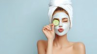 Young woman with white facial mask and towel on head holding cucumber slice over eye for spa skincare treatment on blue background.