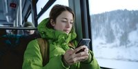 Young Caucasian woman in green winter jacket checking smartphone while traveling by train, snowy landscape visible through window.