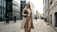 Professional blonde businesswoman in beige coat walking confidently on cobblestone street in modern city center during daytime for corporate lifestyle.