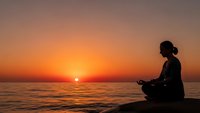 Woman meditating in lotus pose silhouetted against golden sunset over ocean waves. Peaceful mindfulness practice at beach during golden hour.