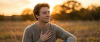Young Caucasian man with reddish-brown hair wearing gray sweater, hand on chest, standing in golden wheat field at sunset. Peaceful, contemplative mood for wellness and lifestyle concepts.
