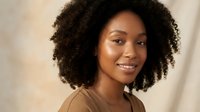 Young African American woman with natural curly hair smiling warmly at camera in casual beige top against soft neutral background for lifestyle content.