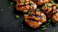 Grilled teriyaki chicken thighs garnished with sesame seeds and chopped green onions on dark background, close up view of glazed Asian style meat.