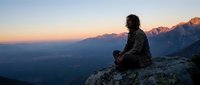 Young woman with long dark curly hair, sitting on mountain rock at sunset, contemplating vast valley landscape below, wearing dark jacket, peaceful meditation moment.