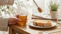 Unrecognizable person holding jar of golden honey with wooden dipper, fresh bread slice and potted herb plant on wooden table.