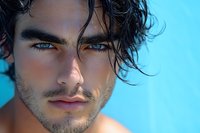 Young Mediterranean male model with intense blue eyes and tousled dark hair against turquoise background, displaying strong facial features and natural stubble.