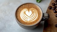 Creamy latte with heart-shaped milk foam art in gray ceramic cup, scattered coffee beans on rustic wooden surface. Close up view with soft natural lighting.