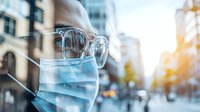 Young Asian woman wearing protective face mask and glasses walking in city street during pandemic, urban background with bokeh sunlight effect.