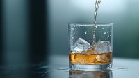 Amber liquid poured into rocks glass with ice cubes, creating dynamic splash and ripples on dark surface against blurred teal background. Perfect for beverage advertising.