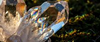 Clear quartz crystal cluster with rainbow light reflections on natural moss surface creating magical prismatic effects in sunlight for healing meditation decor.