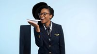 Professional African American female pilot in navy uniform holding paper airplane with confident smile against light blue background.