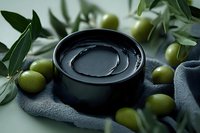 Fresh green olives surround black ceramic bowl with olive oil on gray textile, decorated with olive tree leaves in moody natural lighting for food photography.