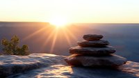 Balanced stone cairn on mountain cliff edge at golden sunrise with dramatic sunburst rays illuminating canyon landscape for meditation and mindfulness concepts.