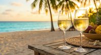 Two glasses of white wine on wooden table at tropical beach sunset with palm trees and turquoise ocean creating romantic vacation atmosphere for couples getaway.