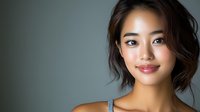 Young Asian woman with natural makeup and gentle smile against dark background, wearing casual top. Professional portrait with soft lighting and shallow depth of field.