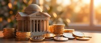 Miniature bank building with classical columns surrounded by stacks of golden coins on wooden surface with warm sunlight creating financial concept.