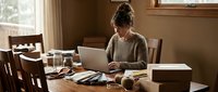 Young woman working from home on laptop at wooden dining table with coffee cup and papers in cozy sunlit room for remote work productivity.