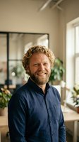 Smiling middle-aged Caucasian businessman with curly hair and beard in navy shirt standing in modern office with plants and natural light.