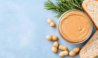 Creamy peanut butter in bowl with whole peanuts, fresh rosemary sprigs and bread slices on light blue background, top view with copy space.