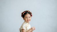 Young Asian girl with braided hair adorned with pink flowers, wearing cream-colored lace top, smiling at camera against white background for children's portraits and family photography.