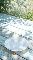Empty transparent plate on weathered wooden table with dappled sunlight and green foliage background, suitable for food styling and outdoor dining concepts.