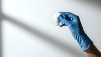 Medical professional hand in blue latex glove holding transparent glass vial for laboratory testing and pharmaceutical research applications.