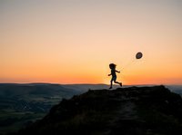 Silhouette of child running on rocky mountain peak at sunset, holding balloon, celebrating achievement and freedom outdoors.
