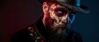 Bearded man with ornate golden skull face paint and cowboy hat in dramatic red and blue lighting for Halloween or Day of the Dead themed projects.