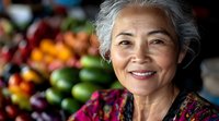 Senior Asian woman smiling warmly at camera in colorful market setting with fresh fruits and vegetables in background for lifestyle and business content.